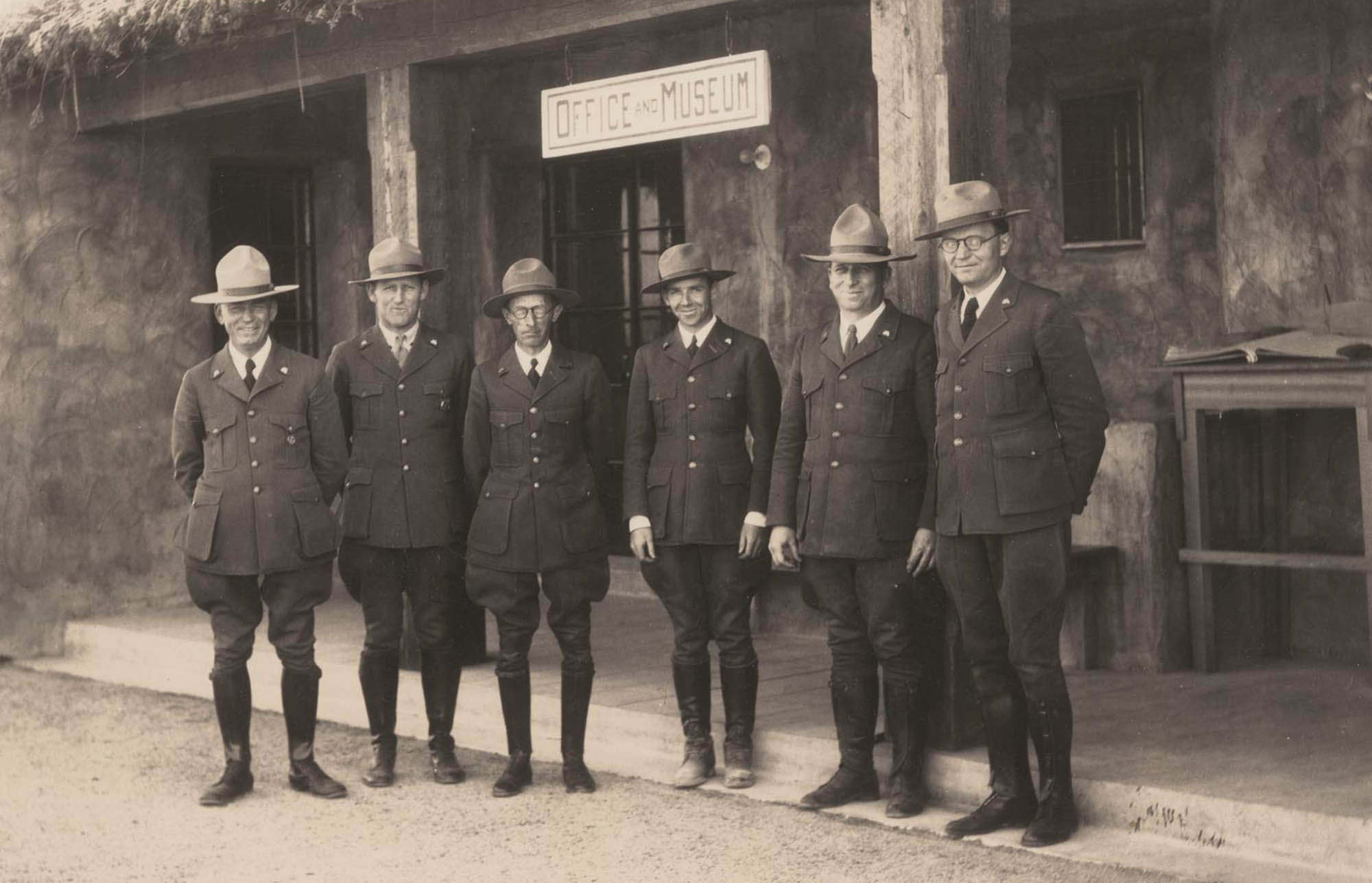 Six men stand in NPS uniforms with broad brim hats. Frank Pinkley wears a round superintendents badge and another ranger wears a shield-shaped badge.